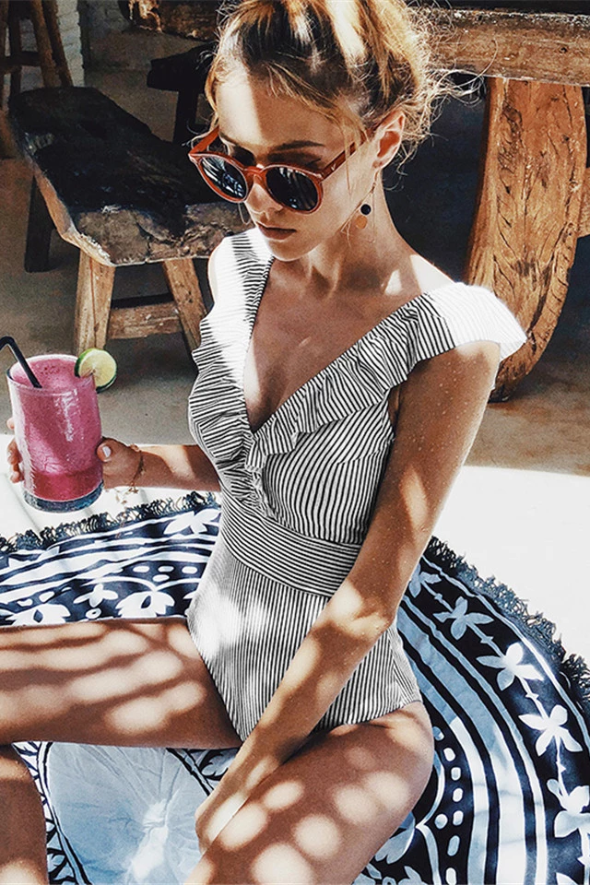 Femme en maillot de bain rayé, lunettes de soleil, sirotant un smoothie rose, assise sur un tapis rond à motifs, ambiance estivale et décontractée.