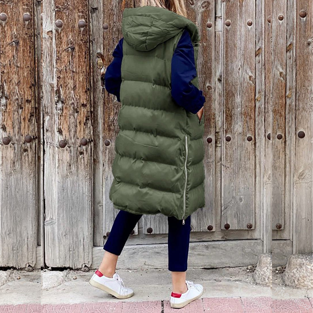 Femme portant un long gilet matelassé vert avec capuche, devant une porte en bois rustique. Mode automne, style décontracté, tendance vestimentaire.