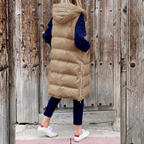 Femme portant un long manteau matelassé beige avec capuche, debout devant une porte en bois rustique. Mode hiver, style décontracté, tendance.