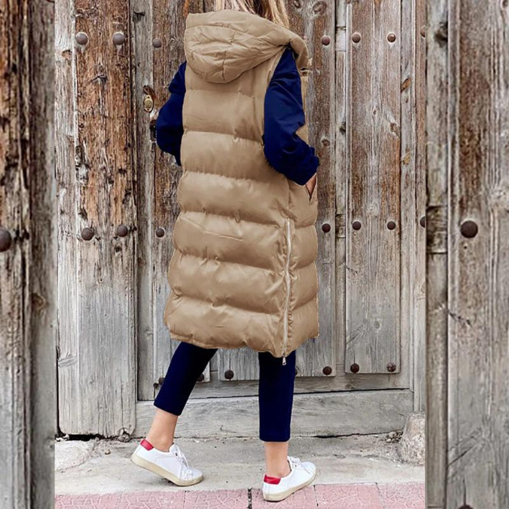 Femme portant un long manteau matelassé beige avec capuche, debout devant une porte en bois rustique. Mode hiver, style décontracté, tendance.