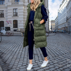 Femme en manteau long vert kaki, tenue décontractée, rue pavée, bâtiments historiques, mode urbaine, sneakers blanches, style automne-hiver.