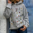 Femme portant un sweat à capuche gris avec motif floral coloré, posant devant une porte bleue. Mode décontractée, tendance automne, style urbain.
