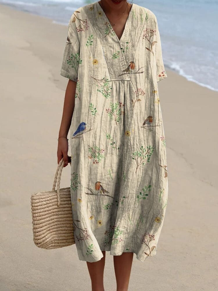 Robe longue en lin beige avec motifs d'oiseaux et de branches, portée par une femme sur la plage, tenant un sac en osier. Mode estivale élégante.