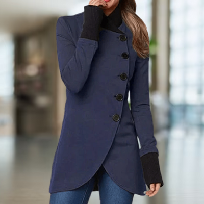 Femme portant un manteau bleu élégant avec boutons noirs, manches longues, col montant, intérieur moderne flou. Mode automne-hiver chic.