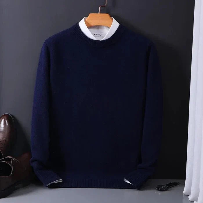 Pull bleu marine en laine sur cintre, col rond, posé contre un mur noir. Chaussures marron et montre noire visibles en arrière-plan.
