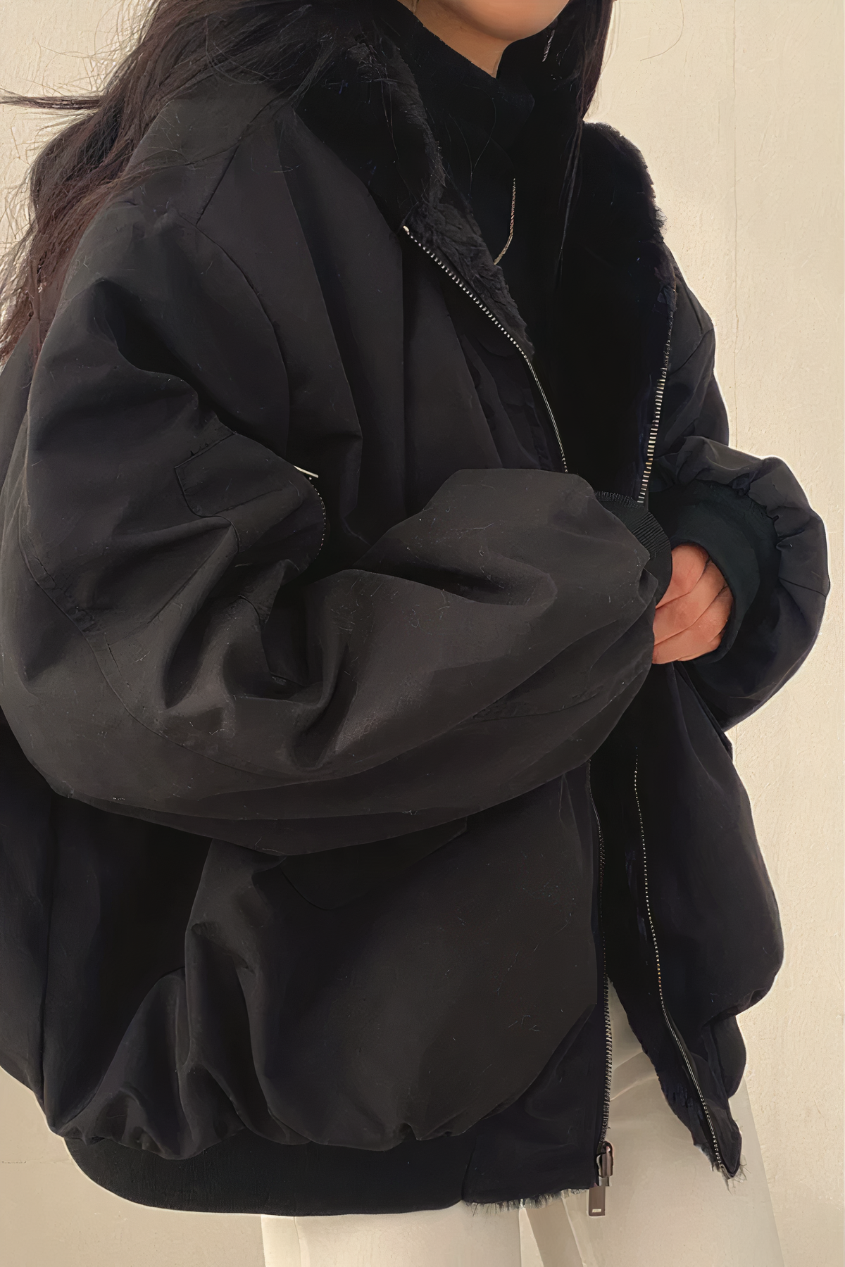 Femme portant une veste noire oversize avec fermeture éclair, manches bouffantes et col en fourrure synthétique, style mode hiver tendance.