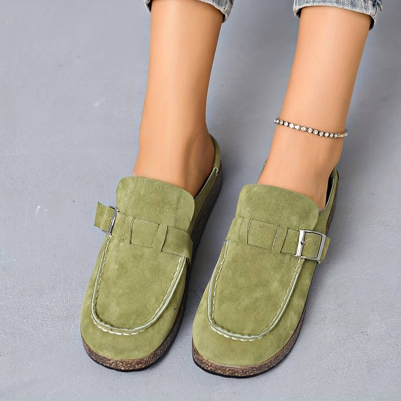 Chaussures mocassins vert olive en daim avec boucle argentée, portées par une personne avec un bracelet de cheville, sur fond gris.