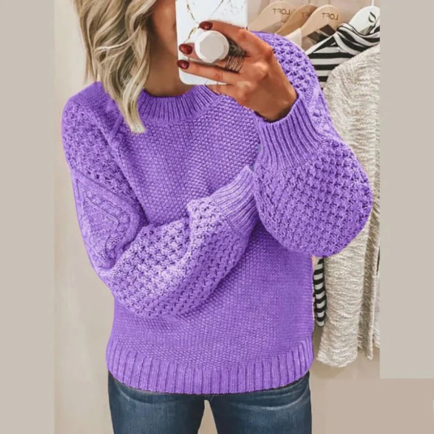 Femme portant un pull violet en tricot torsadé, manches bouffantes, tenant un smartphone, devant un miroir. Mode automne-hiver, style décontracté.