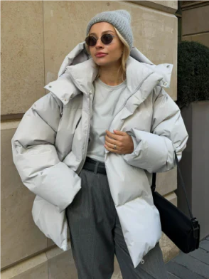 Femme portant une doudoune blanche, bonnet gris et lunettes de soleil, posant devant un mur en pierre. Mode hiver, style urbain chic.
