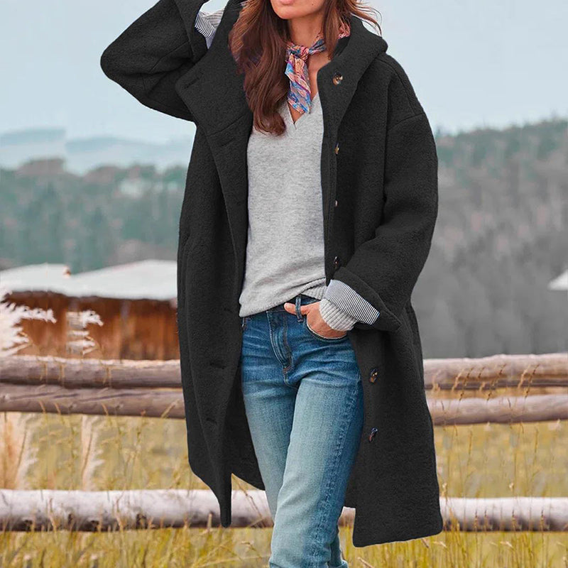 Femme portant un manteau noir élégant, pull gris et jeans, posant en extérieur automnal. Mode féminine, tenue décontractée, style automne-hiver.