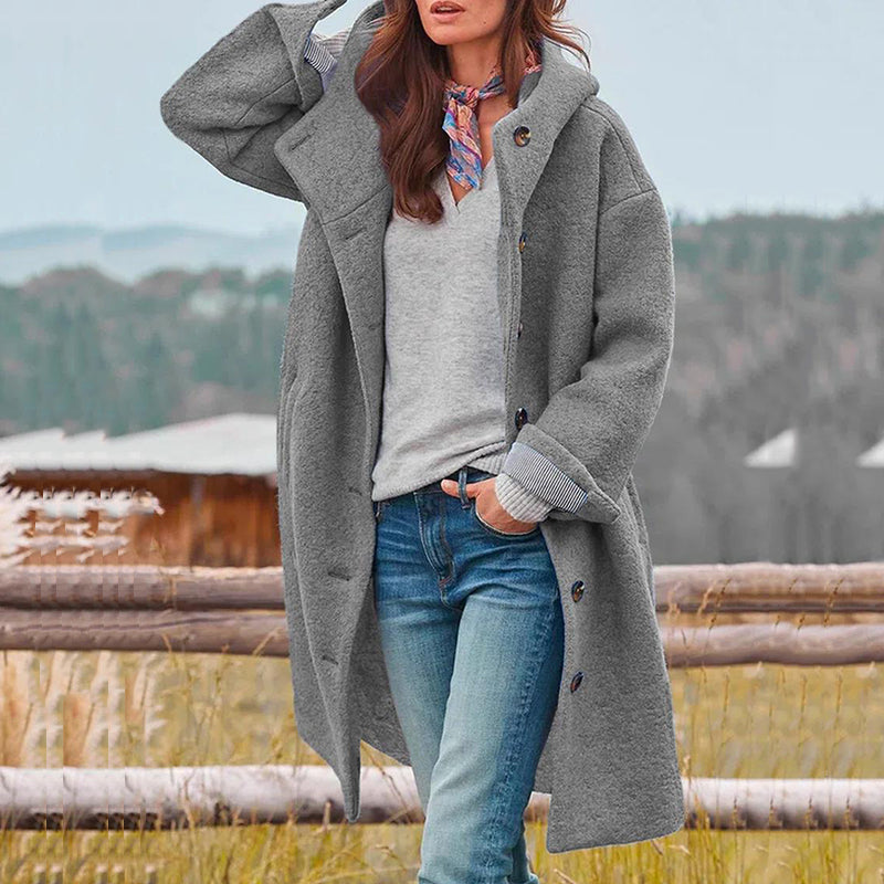 Femme portant un manteau gris en laine, chemise blanche, foulard coloré, posant devant un paysage rural. Mode automne-hiver, style décontracté.