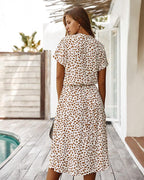 Femme portant une robe à motifs léopard, debout sur une terrasse en bois près d'une piscine, avec une clôture en arrière-plan. Mode été chic.