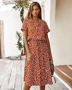 Femme portant une robe rouge à pois blancs, debout sur une terrasse en bois, tenant un sac à main gris. Mode estivale, style décontracté chic.
