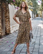 Femme portant une robe imprimé léopard, posant dans une rue pavée bordée de murs en pierre et de fleurs, tenant un sac à main beige.