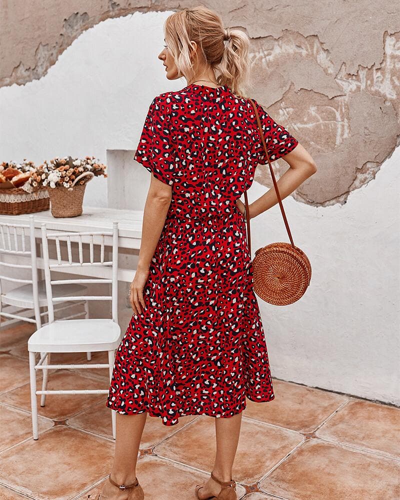 Femme en robe rouge à motifs léopard, sac rond en osier, debout sur terrasse ensoleillée avec chaise blanche et panier de fleurs. Mode estivale chic.