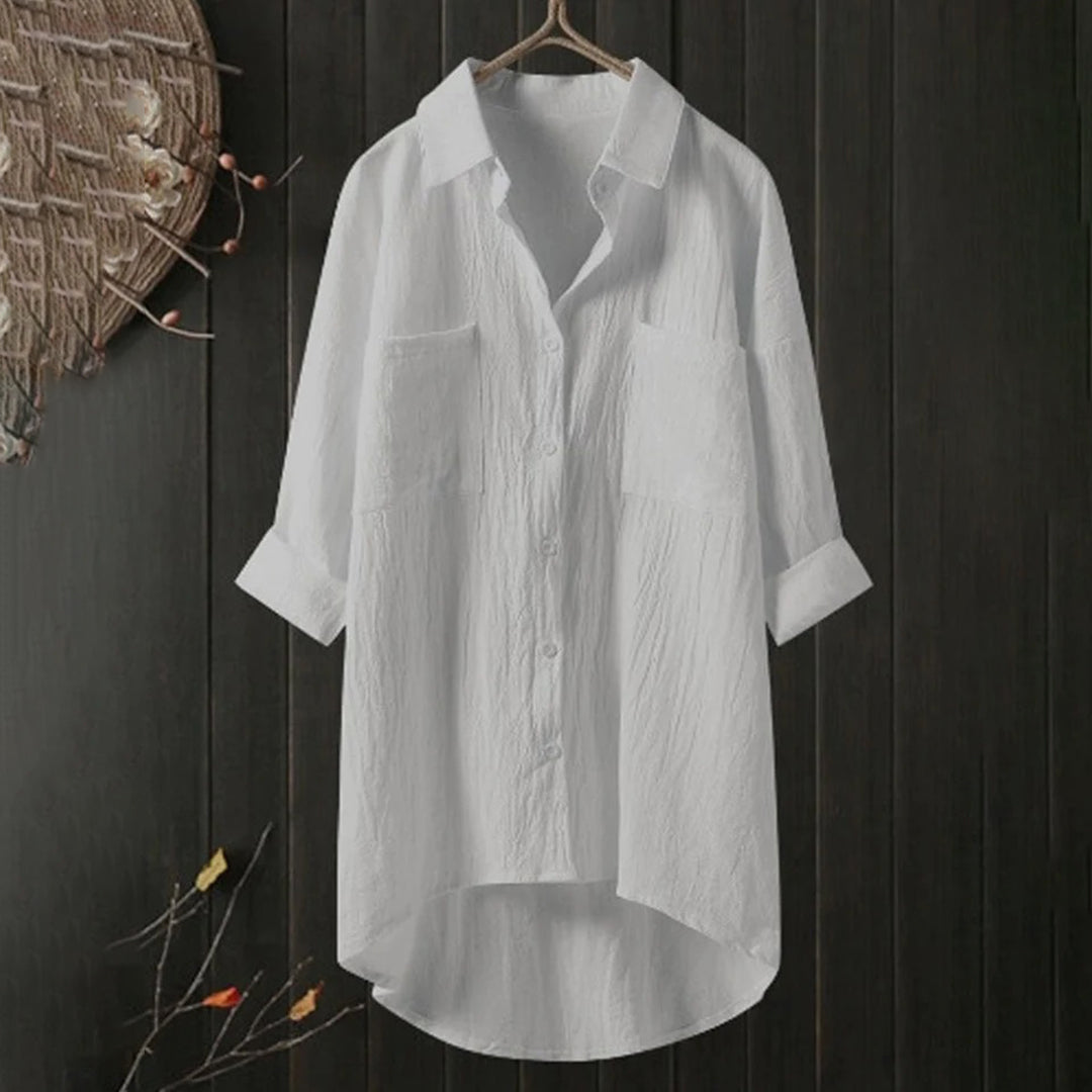 Chemise blanche en lin suspendue sur un cintre, fond noir texturé, manches longues retroussées, col classique, style décontracté et élégant.
