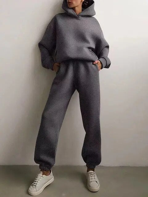 Femme portant un ensemble survêtement gris avec capuche, pantalon ample et baskets blanches. Mode décontractée, tendance automne-hiver.