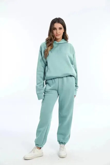 Femme portant un ensemble de survêtement vert menthe, composé d'un sweat à capuche et d'un pantalon, avec des baskets blanches. Mode décontractée.