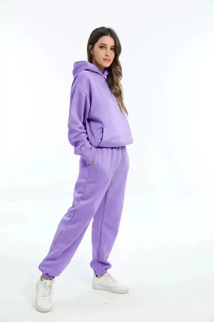 Femme portant un survêtement violet, composé d'un sweat à capuche et d'un pantalon, posant debout. Mode décontractée, tendance streetwear.