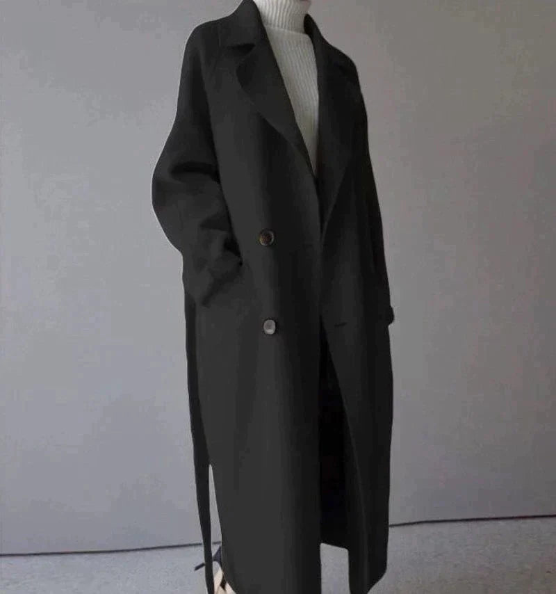 Manteau long noir élégant pour femme, style automne-hiver, avec boutons et poches, porté sur un pull blanc, mode chic et moderne.