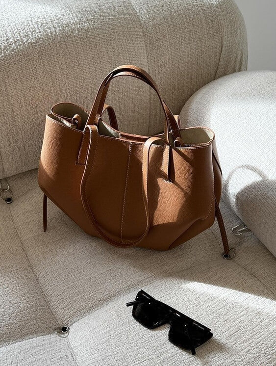 Sac à main en cuir marron sur un canapé beige, accompagné de lunettes de soleil noires. Accessoire mode élégant et tendance pour femme.