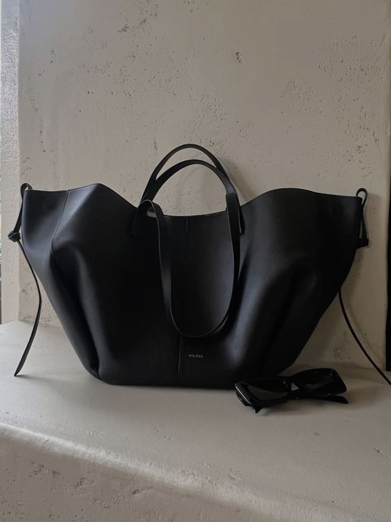 Sac à main noir en cuir élégant posé sur une surface blanche, accompagné de lunettes de soleil noires. Accessoire mode chic et tendance.