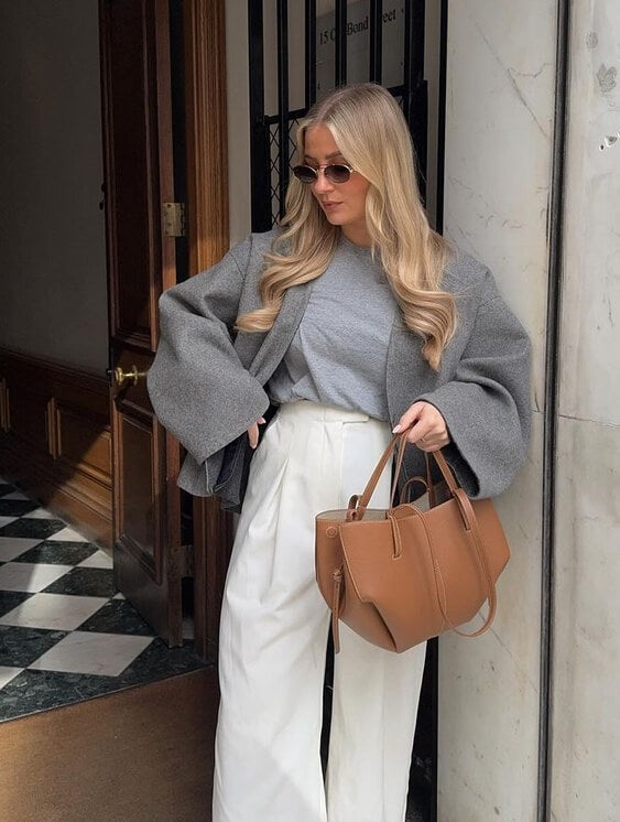 Femme élégante portant un manteau gris, pantalon blanc et lunettes de soleil, tenant un sac à main marron. Mode automne chic, style urbain tendance.