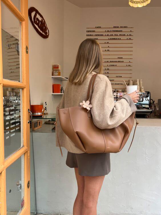 Femme de dos dans un café, portant un grand sac en cuir marron et tenant un gobelet de café. Menu visible en arrière-plan. Ambiance décontractée.