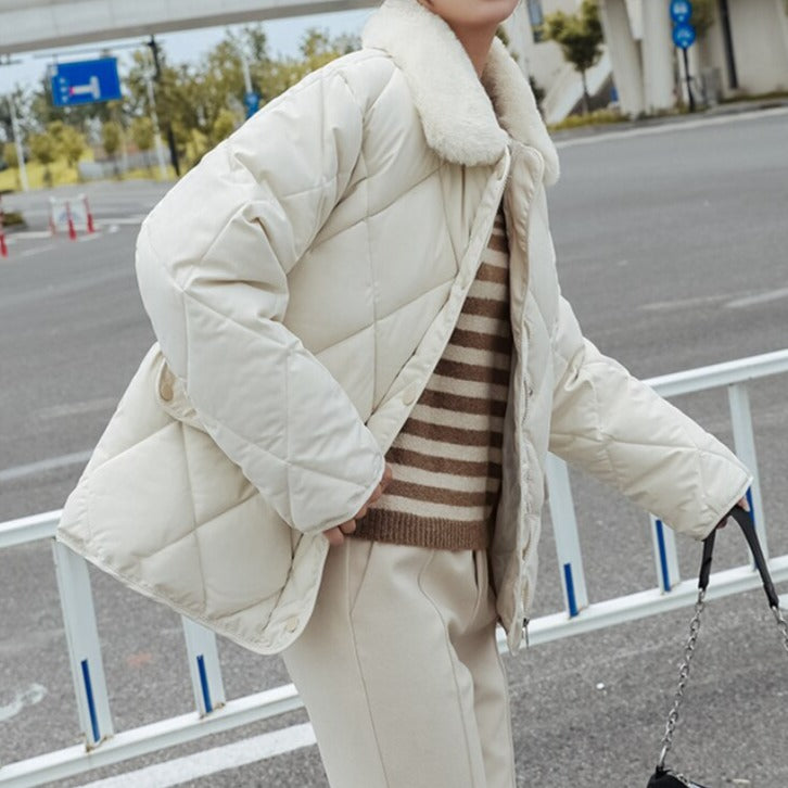 Veste matelassée blanche avec col en fourrure, portée par une personne en pull rayé et pantalon beige, sur fond de rue urbaine.