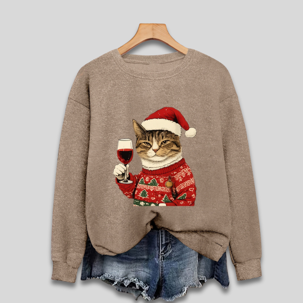 Pull beige avec motif de chat portant un bonnet de Noël, tenant un verre de vin rouge, sur cintre en bois. Mode festive, style décontracté.