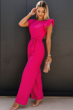 Femme en combinaison rose vif, posant contre un mur noir, tenant une pochette beige. Mode élégante, tenue chic, style moderne.