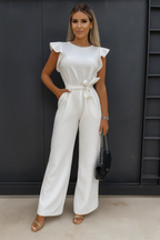 Femme en combinaison blanche élégante avec ceinture nouée, posant devant un mur sombre. Mode féminine chic, tenue de soirée tendance.