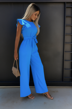 Femme en combinaison bleue élégante, tenant un sac à main beige, posant devant un mur noir. Mode féminine, style chic, tenue tendance.