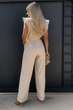 Femme en combinaison beige élégante, vue de dos, devant une porte noire moderne. Mode chic, cheveux longs blonds, talons hauts.