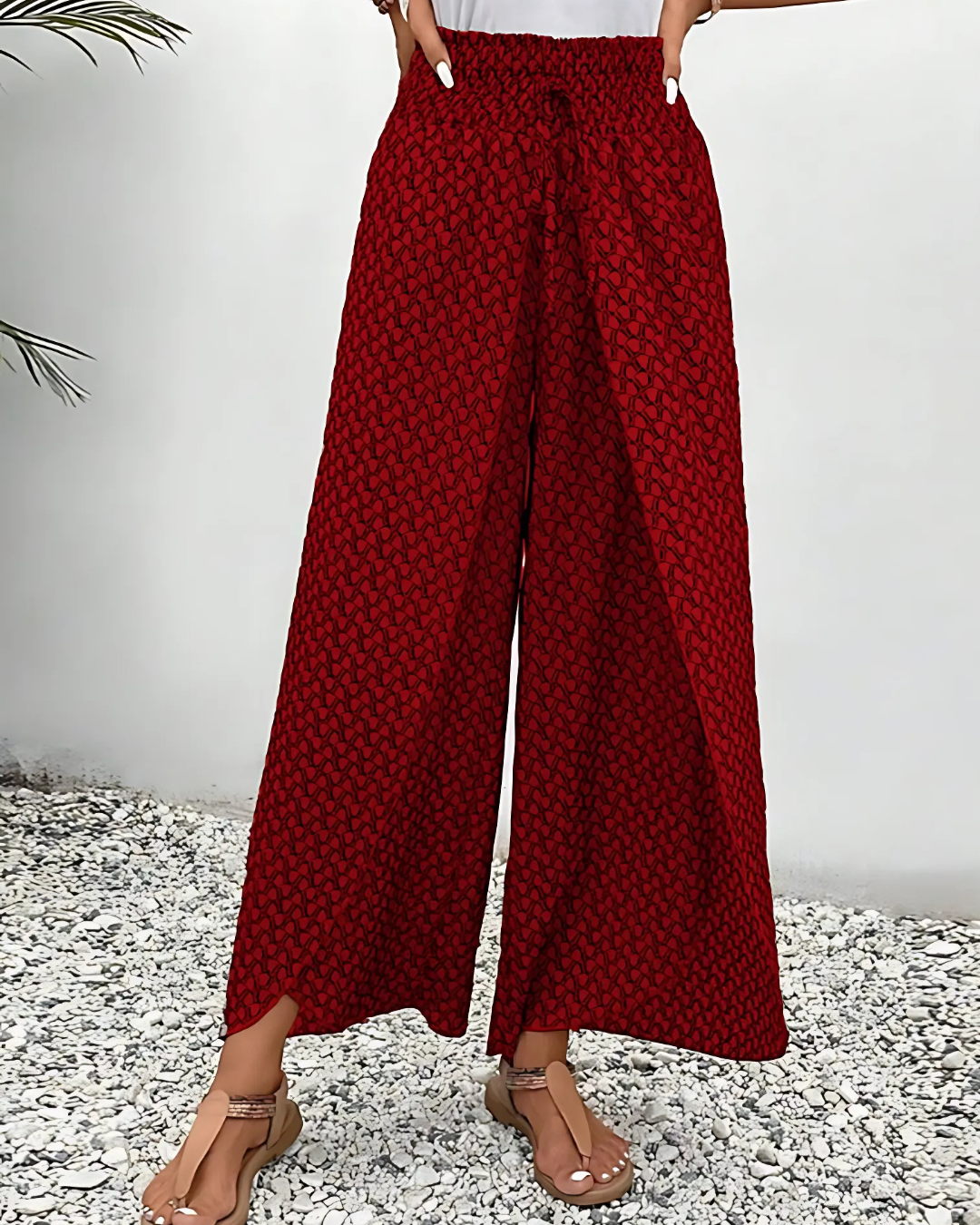 Pantalon large rouge à motifs géométriques noirs, style bohème, porté par une femme sur fond de gravier blanc. Mode estivale décontractée.