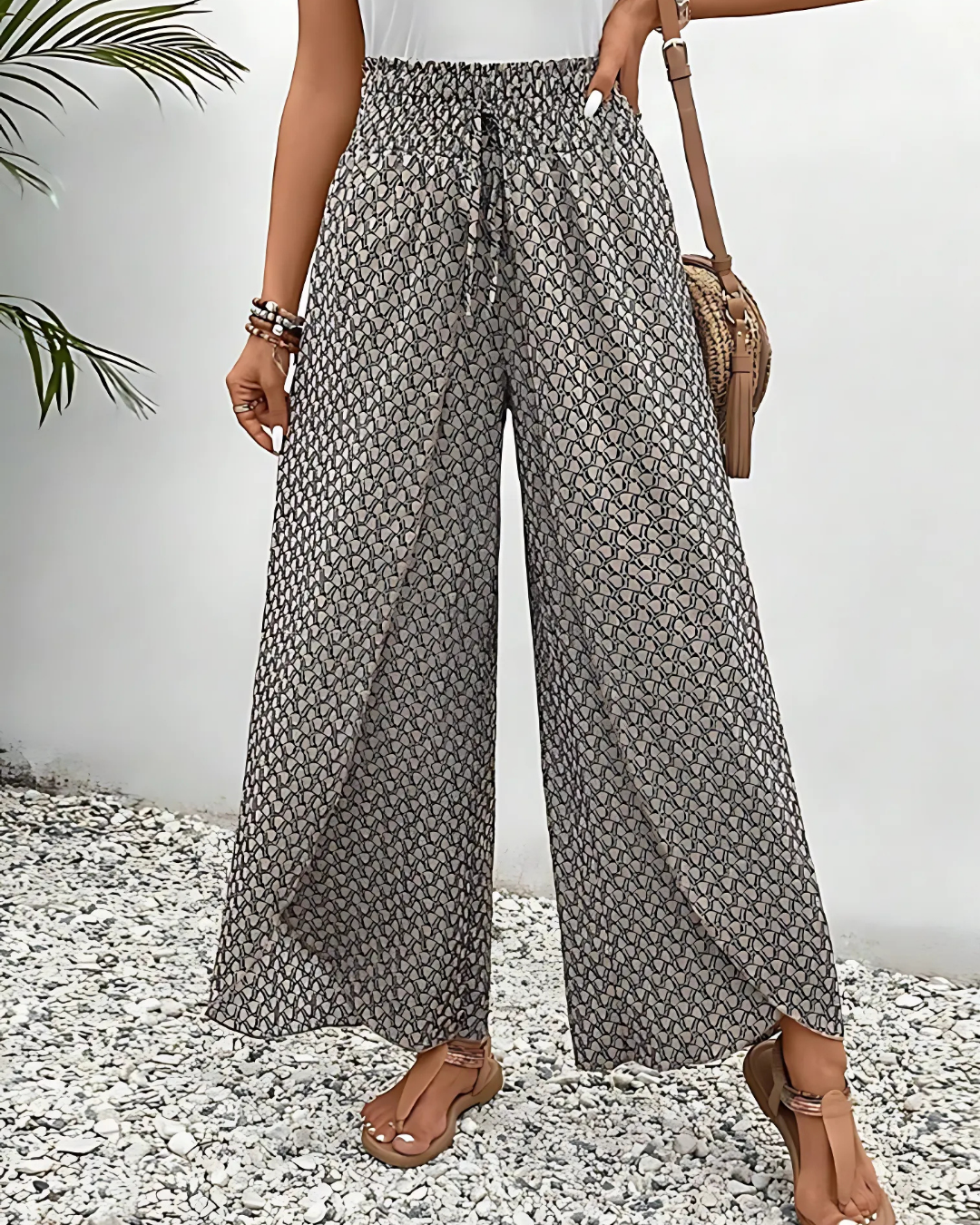 Pantalon large femme à motifs géométriques, taille haute élastique, porté avec un sac en bandoulière marron et sandales, sur fond de gravier.