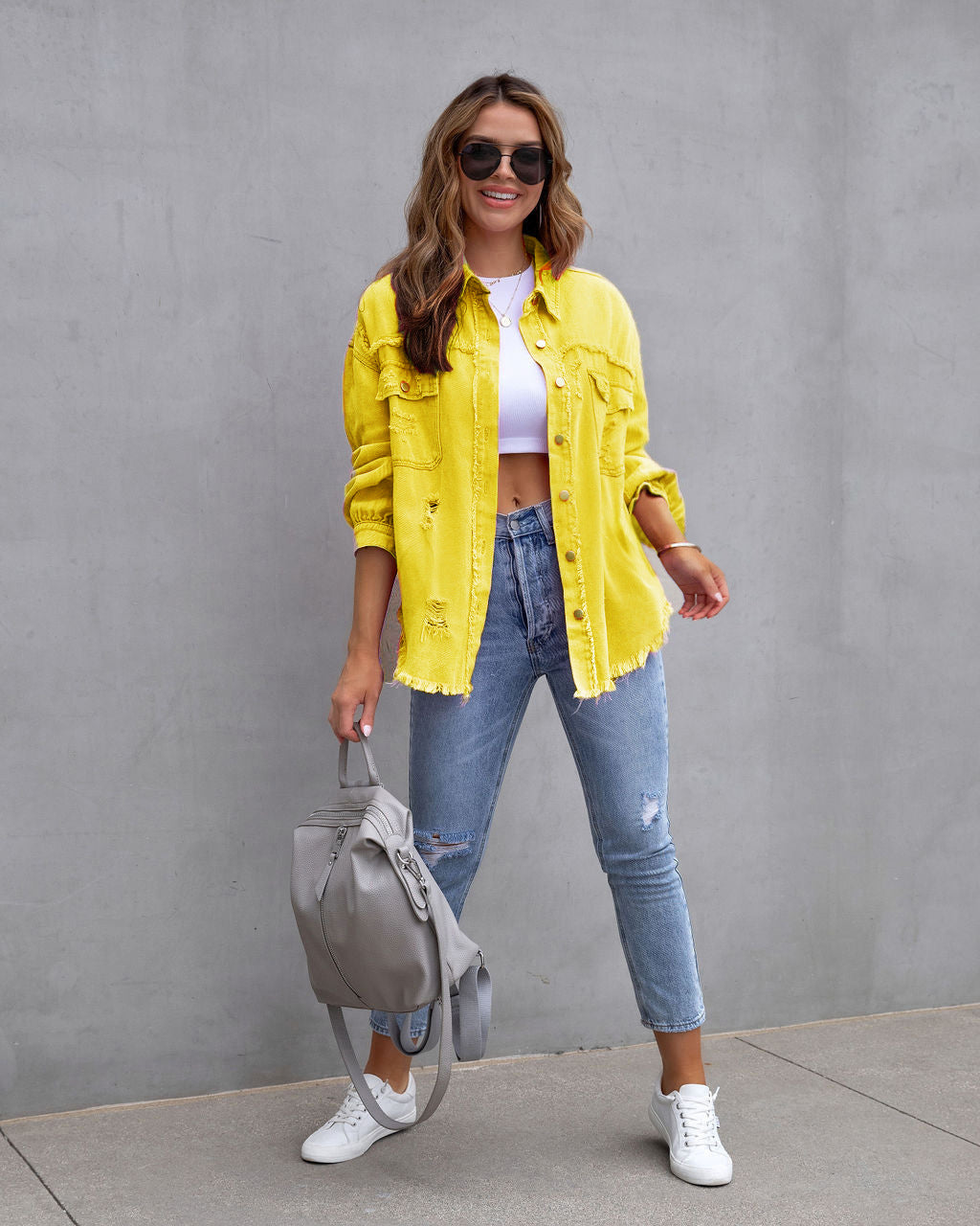 Femme souriante en chemise jaune, jean déchiré, baskets blanches, portant un sac à dos gris, posant devant un mur gris. Mode décontractée tendance.