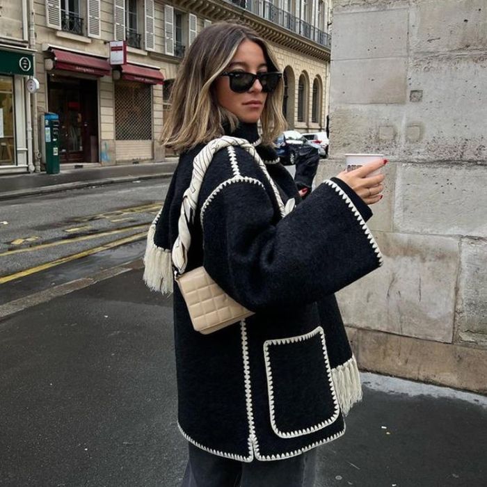 Jeune femme portant un manteau noir avec bordures blanches, lunettes de soleil, sac beige, tenant un café, dans une rue parisienne.