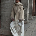 Femme élégante en manteau beige et écharpe assortie, pantalon blanc ample, tenant un sac tissé, posant devant un mur en pierre. Mode automne chic.