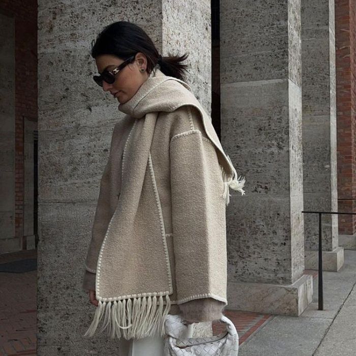 Femme portant un manteau beige avec écharpe assortie, lunettes de soleil, sac blanc tressé, devant un mur en pierre. Mode automne-hiver élégante.