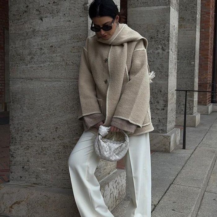 Femme en manteau beige élégant, pantalon blanc, lunettes de soleil, tenant un sac à main tressé blanc, posant devant un mur en pierre.