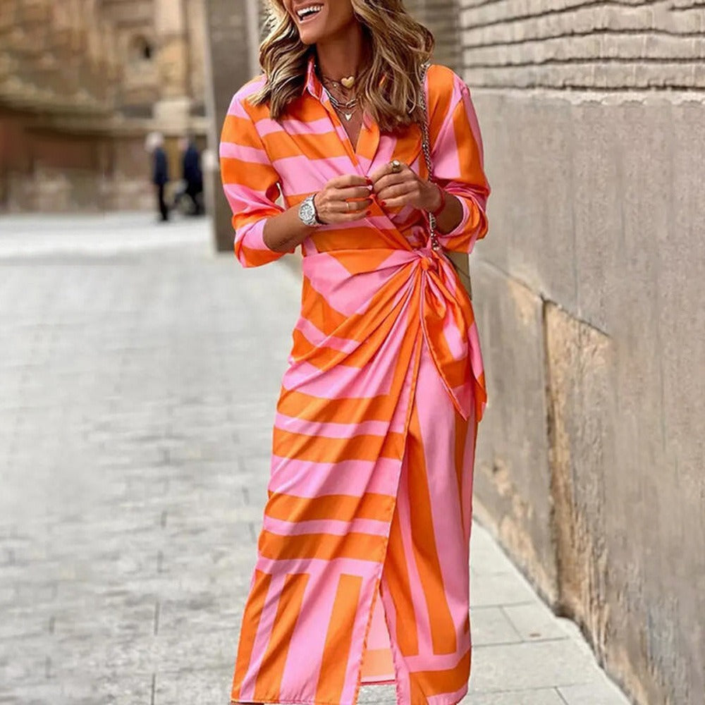 Robe longue rayée rose et orange, manches longues, portée par une femme souriante dans une rue pavée. Mode élégante, tendance automne.