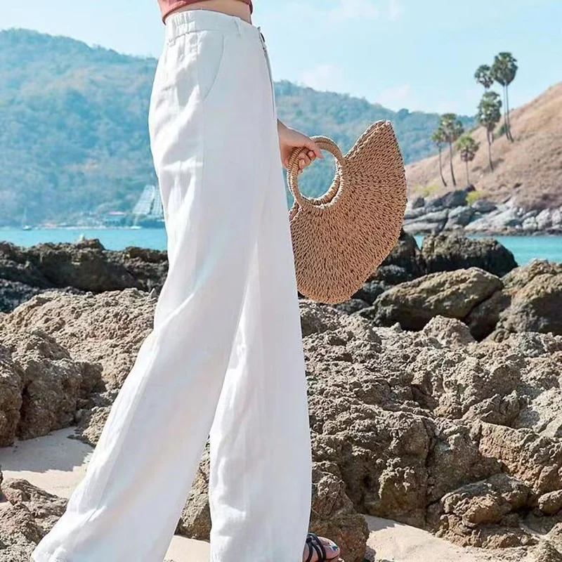 Femme en pantalon blanc sur plage rocheuse, tenant un sac en paille. Paysage côtier en arrière-plan avec mer et collines. Mode estivale chic.
