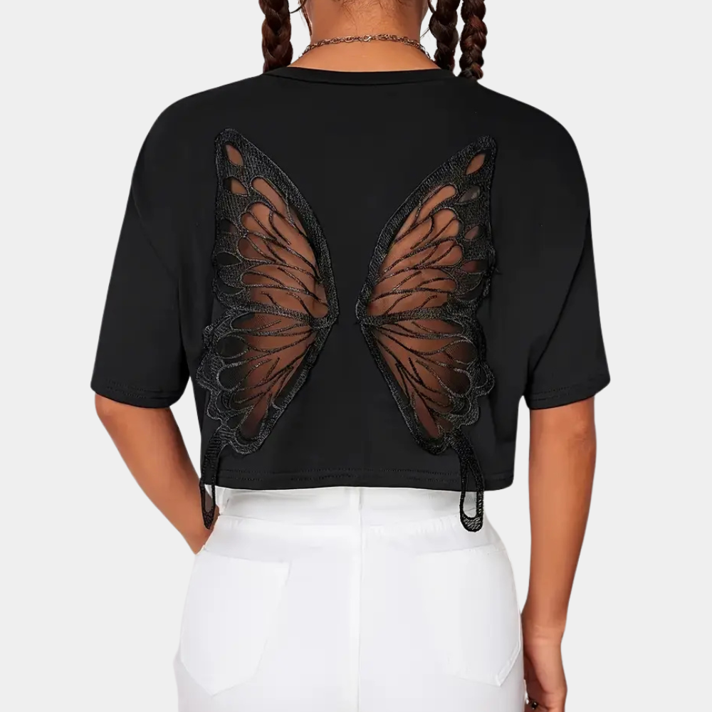 Femme portant un t-shirt noir avec motif ailes de papillon en dentelle au dos, coiffure tresses, pantalon blanc. Mode tendance, style unique.