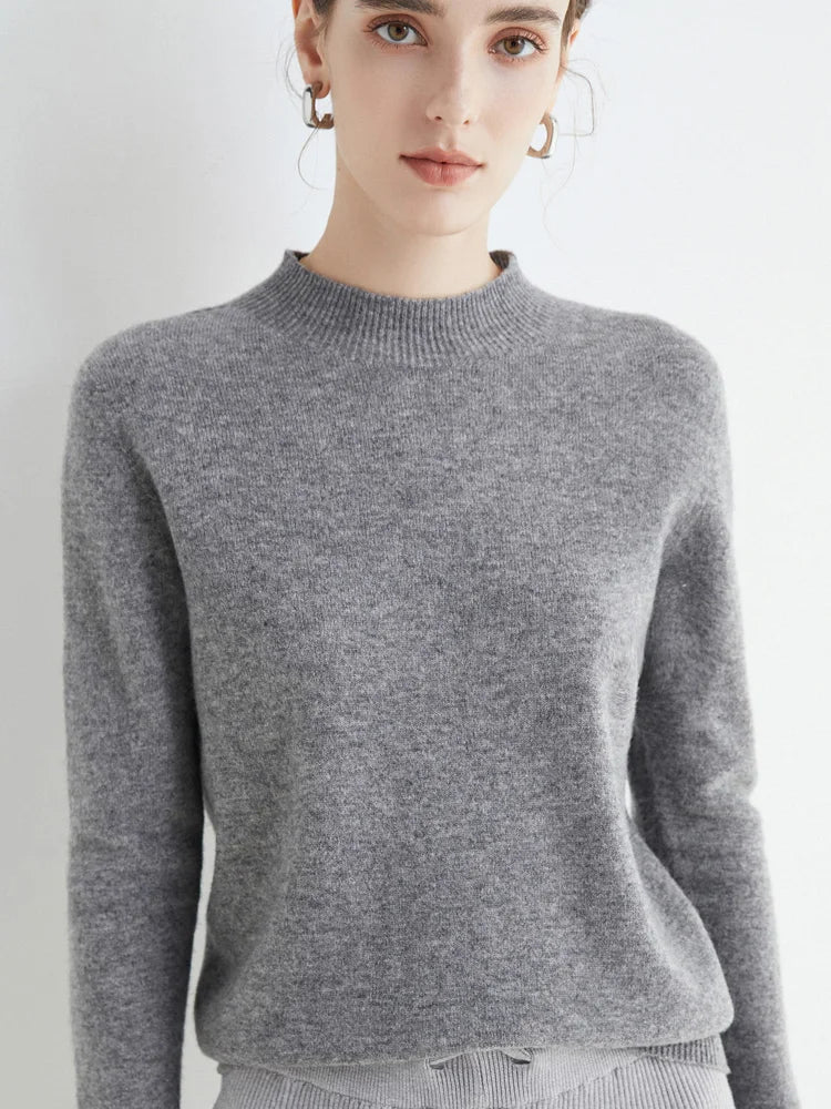 Femme portant un pull gris en cachemire, manches longues, col rond, style élégant et minimaliste, mode automne-hiver, fond blanc.