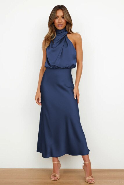 Robe longue bleu marine en satin, col drapé, sans manches, portée par une femme debout sur un sol en bois, élégance et style moderne.