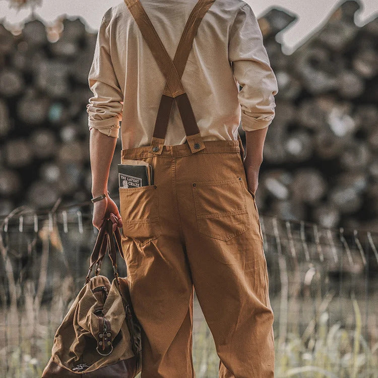 Homme en salopette marron tenant un sac en toile, livre dans la poche arrière, fond de bûches flou. Mode rétro, style décontracté, nature.