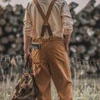 Homme en salopette marron tenant un sac en toile, livre dans la poche arrière, fond de bûches flou. Mode rétro, style décontracté, nature.