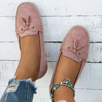 Mocassins roses en daim pour femme, avec nœuds décoratifs, portés avec un jean déchiré et un bracelet de cheville perlé, sur fond de bois blanc.