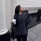 Femme élégante tenant un gobelet de café, portant un manteau gris, posant devant une clôture en fer forgé. Mode urbaine, style automnal.